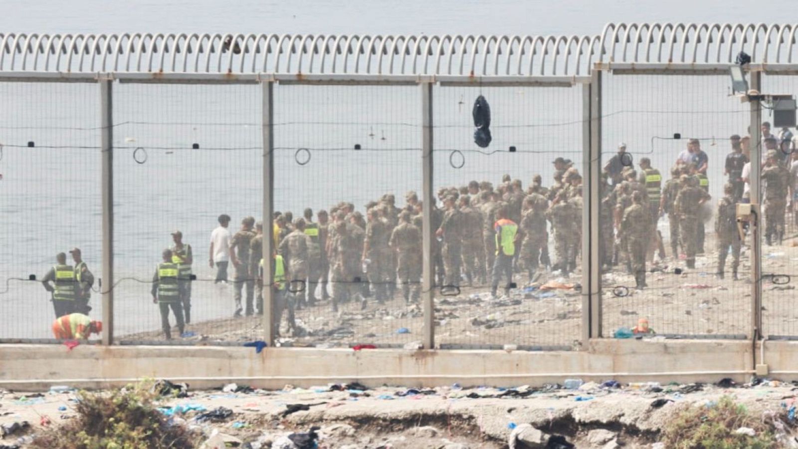 Valla fronteriza en el Tarajal con basura en Ceuta y grupo de personas uniformadas en Marruecos. Una persona con chaleco naranja en primer plano.