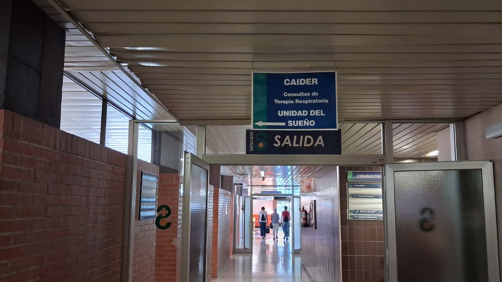 Un pasillo de la Unidad de Neumología del Hospital Universitario de Guadalajara con tres personas andando al fondo y un cartel azul y verde que dirige hacia la Unidad del Sueño. Bajo este aparece otro cartel gris en el que se lee "SALIDA".