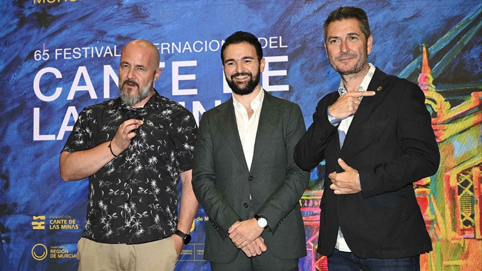 Joaquín Zapata, Ricardo Castella y Carlos del Amor en el 65 Festival Internacional del Cante de las Minas. Zapata con camisa de palmeras, Castella con traje verde, y Del Amor señalando. Fondo azul con texto del festival y logos.
