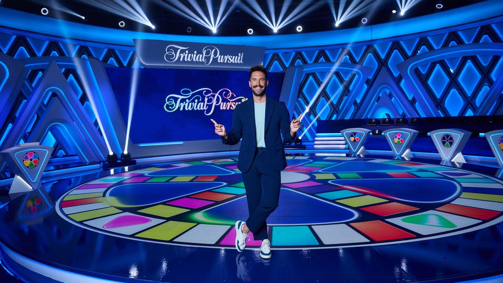 Egoitz Txurruka "Txurru" en el plató de "Trivial Pursuit", con traje azul, camiseta clara y zapatillas. Pose dinámica con brazos levantados y dedos apuntando. Fondo azul con logo del programa, tablero circular y podios.