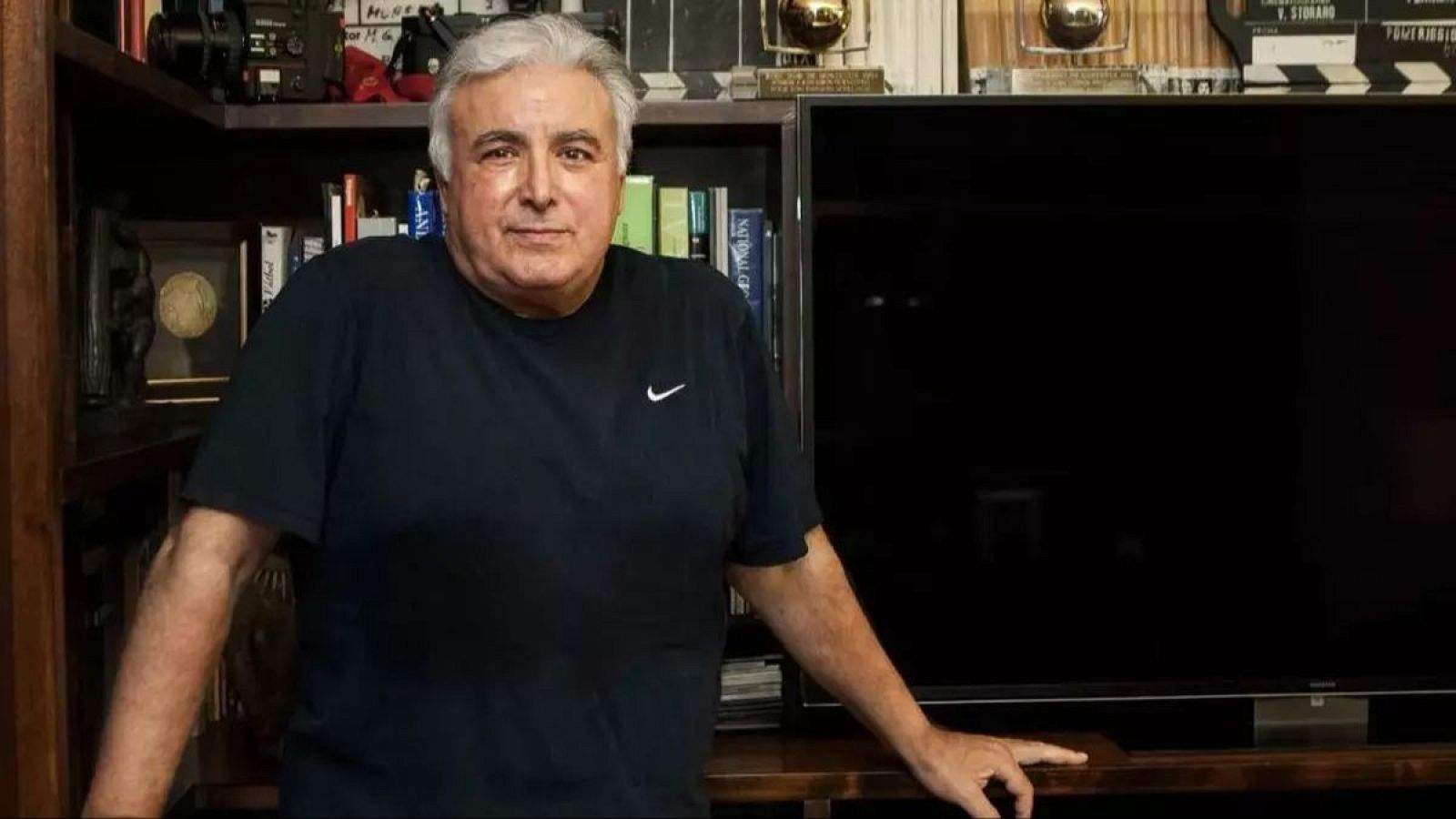 Retrato de Juan Lebrón, productor y cineasta, en su estudio. Luce una camiseta negra de Nike y posa frente a una estantería con premios y una claqueta.