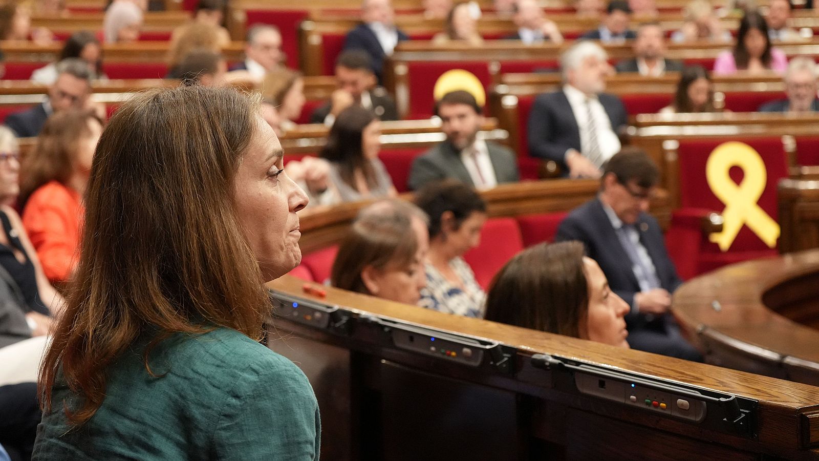 Jéssica Albiach, presidenta de los Comuns en el Parlament, de perfil derecho, con camisa verde azulado y expresión seria, en el hemiciclo tras una votación. Se aprecia un lazo amarillo en el fondo.