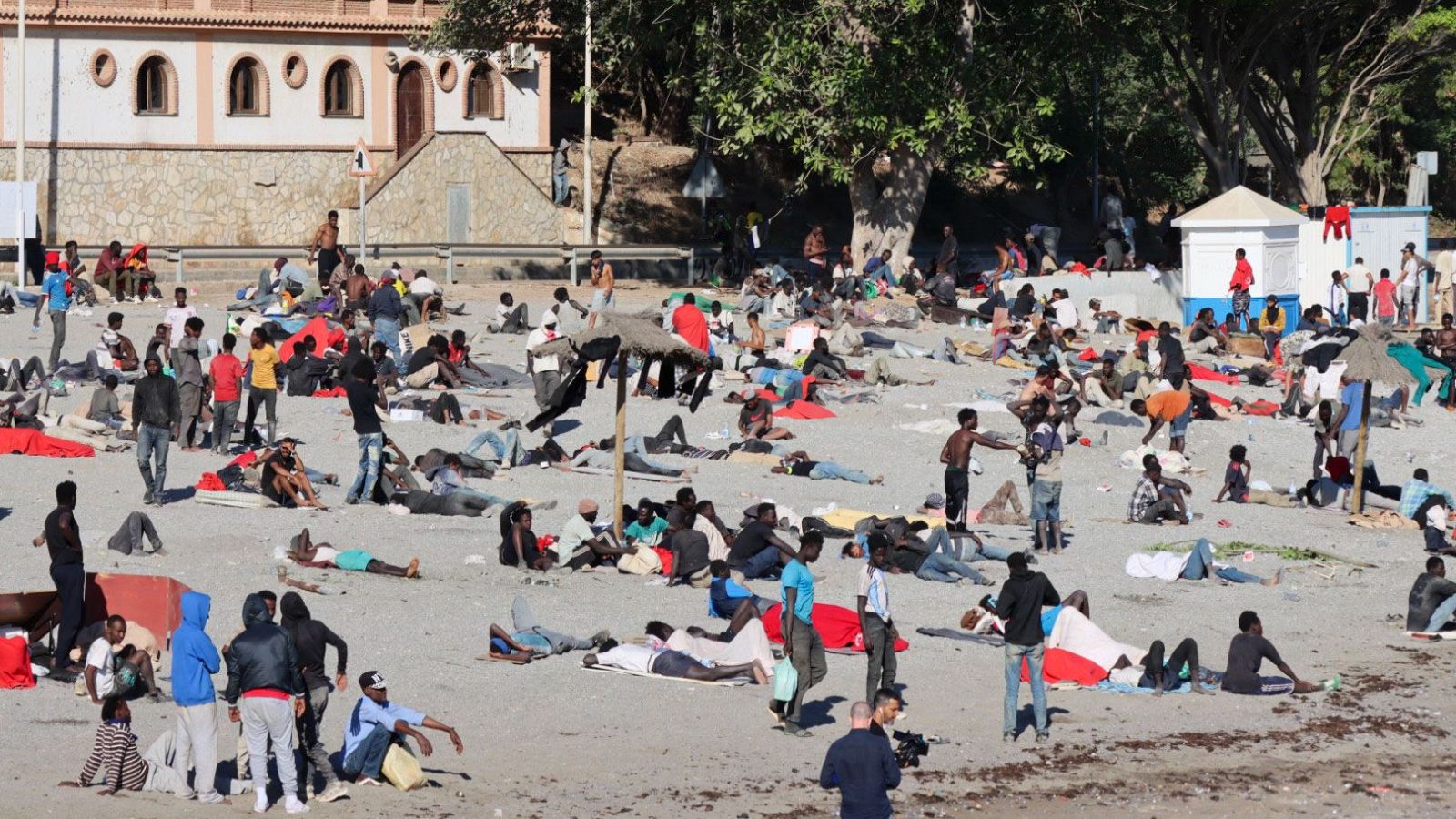 Vista aérea de la playa 'El Trampolín' en Ceuta, con cientos de personas, mayormente hombres jóvenes, descansando sobre la arena con mantas de colores. Al fondo, un edificio y vegetación.