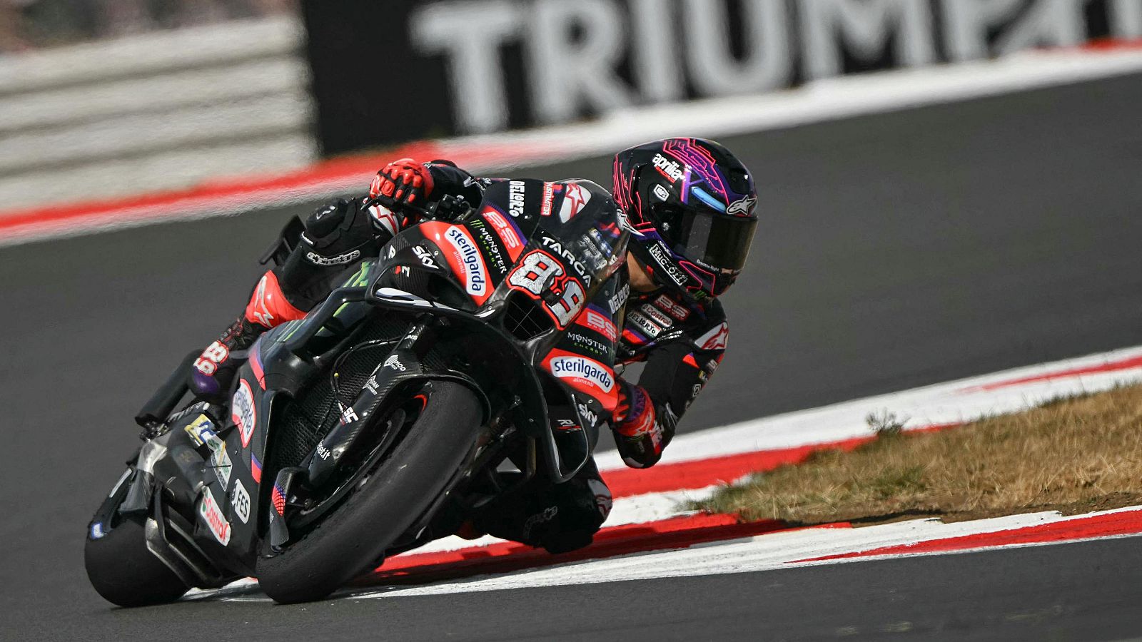 Jorge Martín (89) en acción en un circuito de MotoGP, inclinado en una curva sobre su motocicleta negra con detalles rojos y blancos. Su casco presenta un diseño geométrico llamativo.