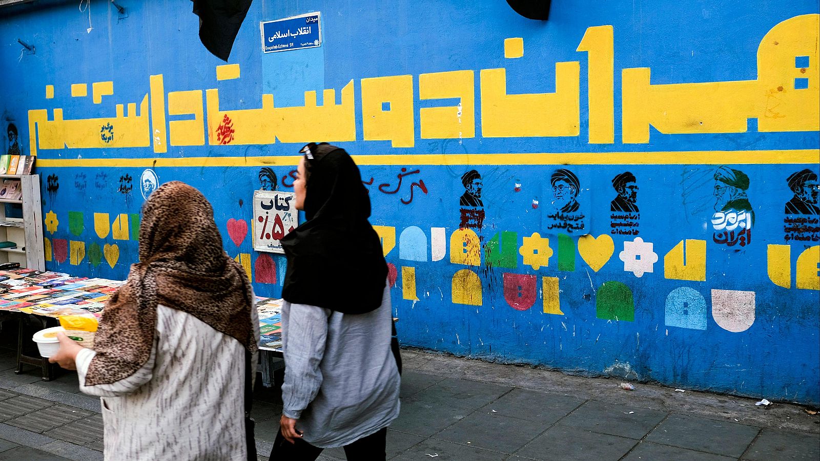 Mural azul en Irán con texto persa, retratos de hombres con turbantes y símbolos coloridos. Dos mujeres caminan en primer plano. Puesto de libros a la izquierda.