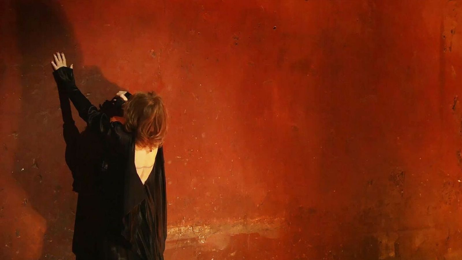 Fotograma de 'Seize moments de ma vie' de Albert Serra. Persona de espaldas con brazos extendidos hacia pared rojiza rugosa. Iluminación dramática.