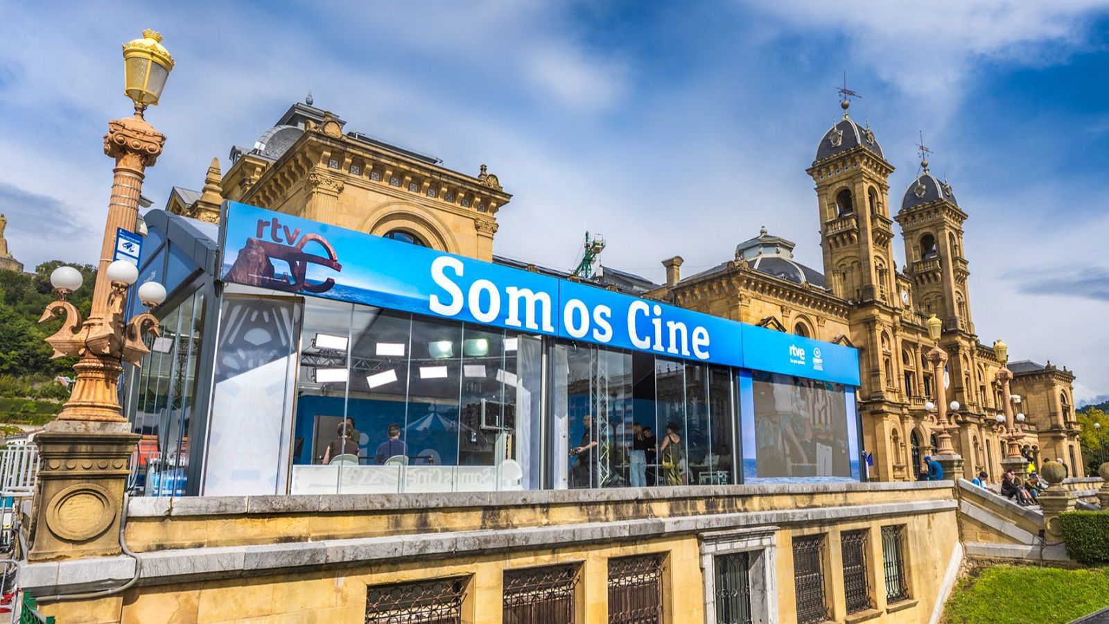 Pabellón "Somos Cine" de RTVE frente al Kursaal de San Sebastián, con el "Peine del Viento" y personas en su interior. Cielo azul y farola clásica a la izquierda.
