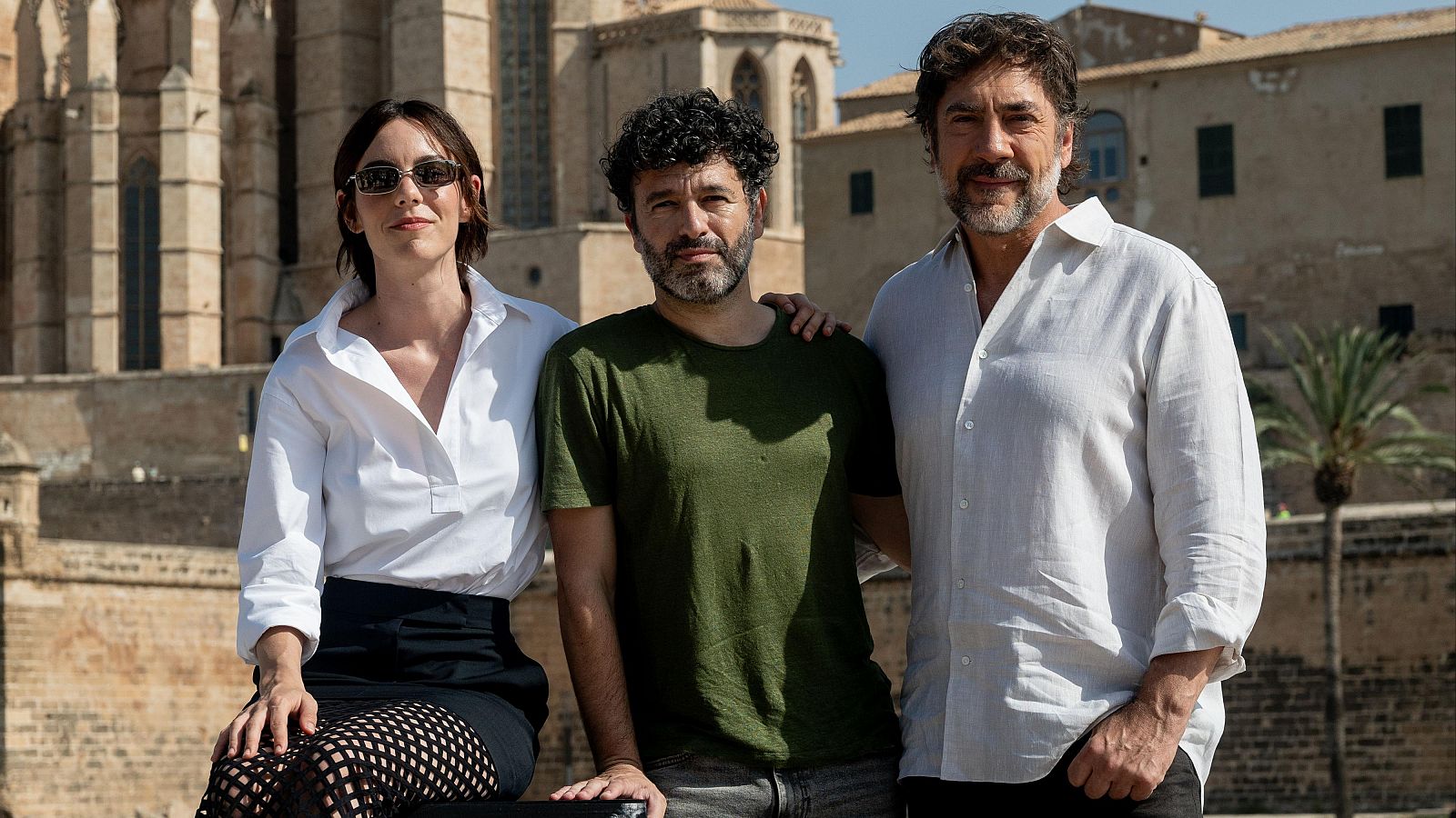 Victoria Luengo, Rodrigo Sorogoyen y Javier Bardem posan juntos al aire libre con un edificio histórico y una palmera de fondo. Luengo lleva gafas de sol, camisa blanca y falda oscura con medias de rejilla.