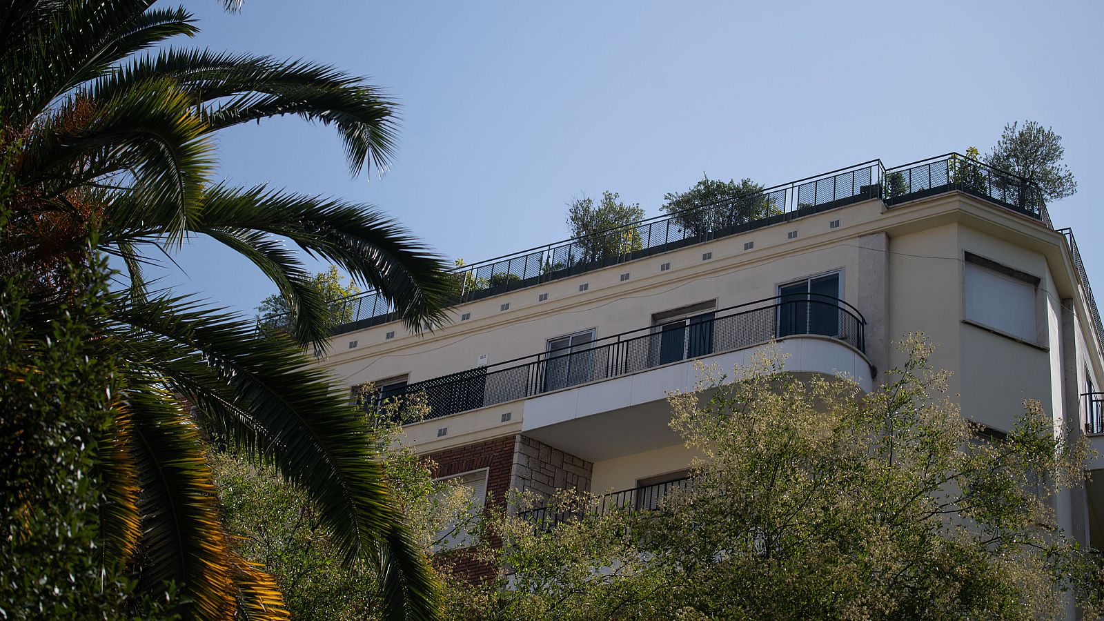 Fachada de edificio residencial moderno con azotea ajardinada, balcones y vegetación, bajo un cielo azul.