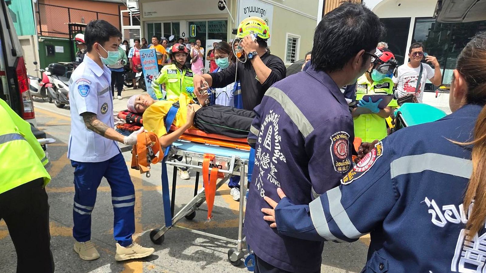 Rescatistas tailandeses trasladan a un herido en camilla, con detalles de uniformes y texto en tailandés visible. La escena transcurre en un entorno urbano con edificios y una ambulancia al fondo.