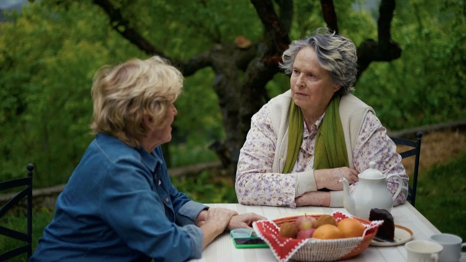 Mercedes Milá y Lucía Dominguín conversan en un jardín, con una mesa llena de frutas, pastel y teteras. Un día soleado y un entorno natural las acompañan.