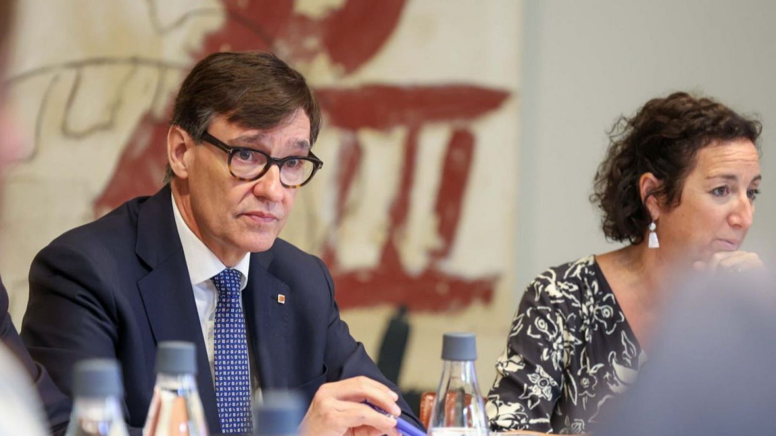 Primer plano de Salvador Illa en una reunión, con traje oscuro y corbata azul, junto a una mujer con pendientes largos y blancos. Botellas de agua y arte abstracto al fondo.