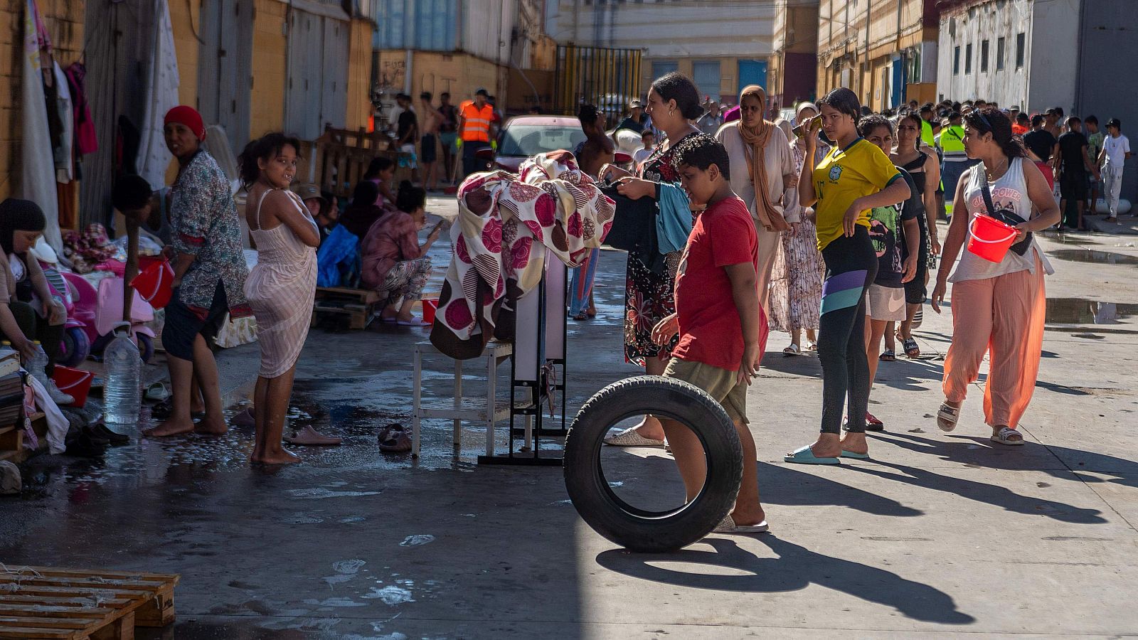 Calle concurrida en Ceuta, personas esperando bajo el sol, suelo mojado, botellas de agua, palés y ropa tendida. Se aprecian chalecos de alta visibilidad y edificios al fondo.