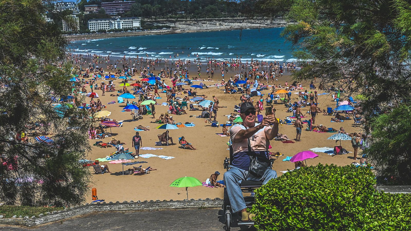Hombre en silla de ruedas se toma un selfie en la playa del Sardinero, Santander, con bañistas y sombrillas de colores de fondo.
