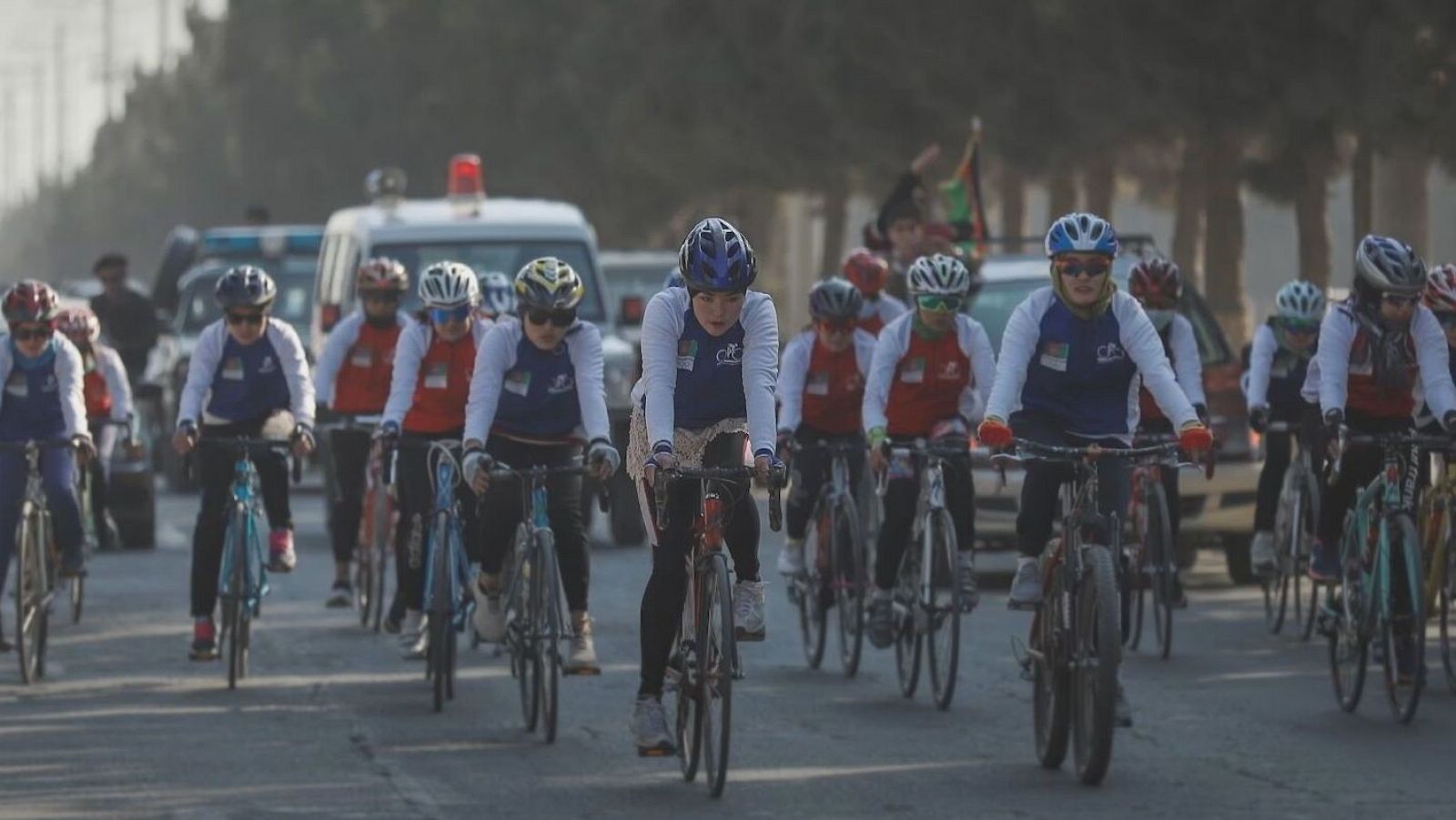 Grupo de ciclistas, predominantemente mujeres, en plena competición sobre una carretera. Destaca una ciclista con maillot blanco y azul, casco azul y bicicleta roja.