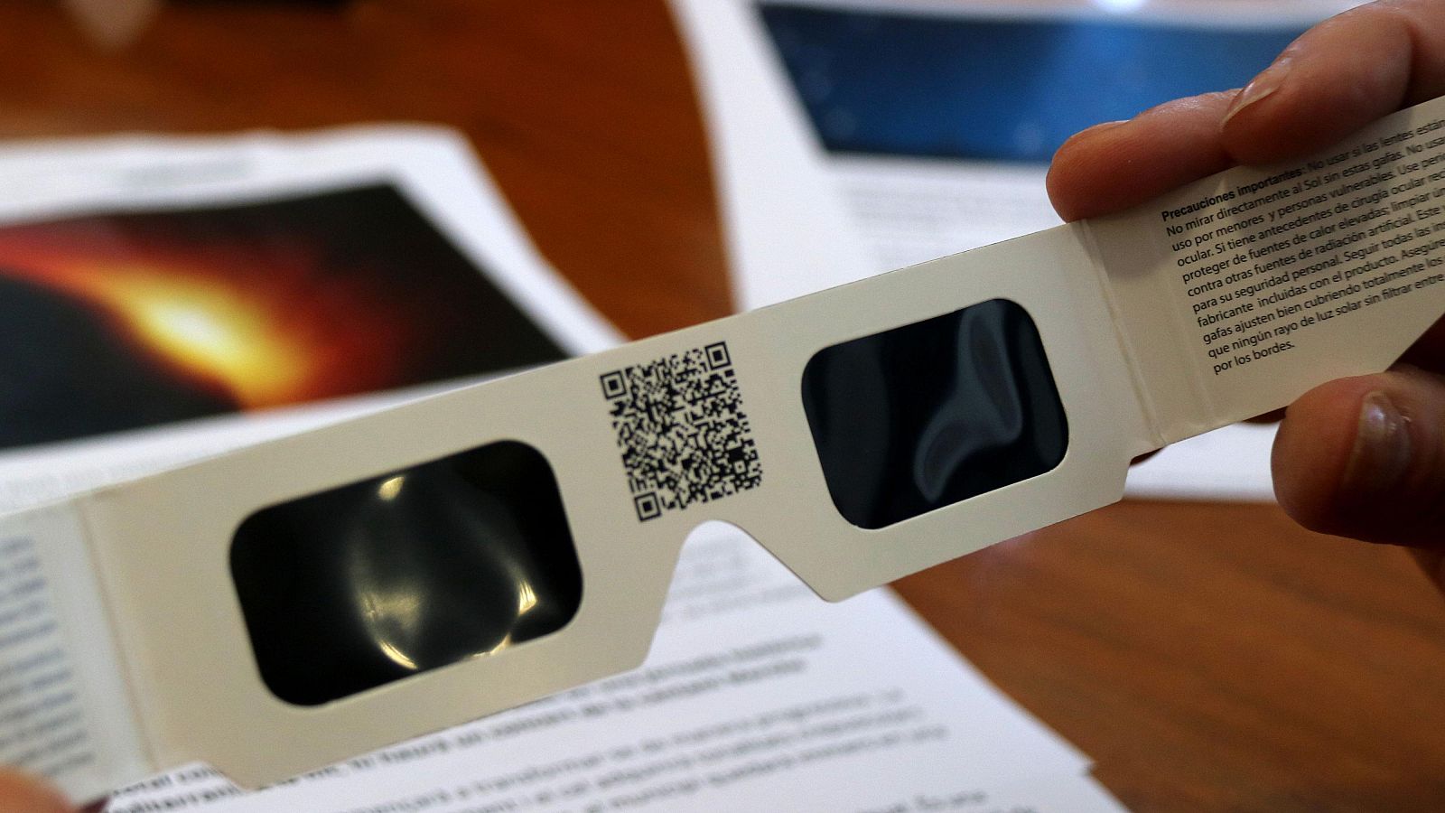 Gafas de eclipse de cartón blanco con lentes oscuras, código QR y advertencias de seguridad impresas: "Precauciones importantes", "No mirar directamente al sol sin estas gafas".