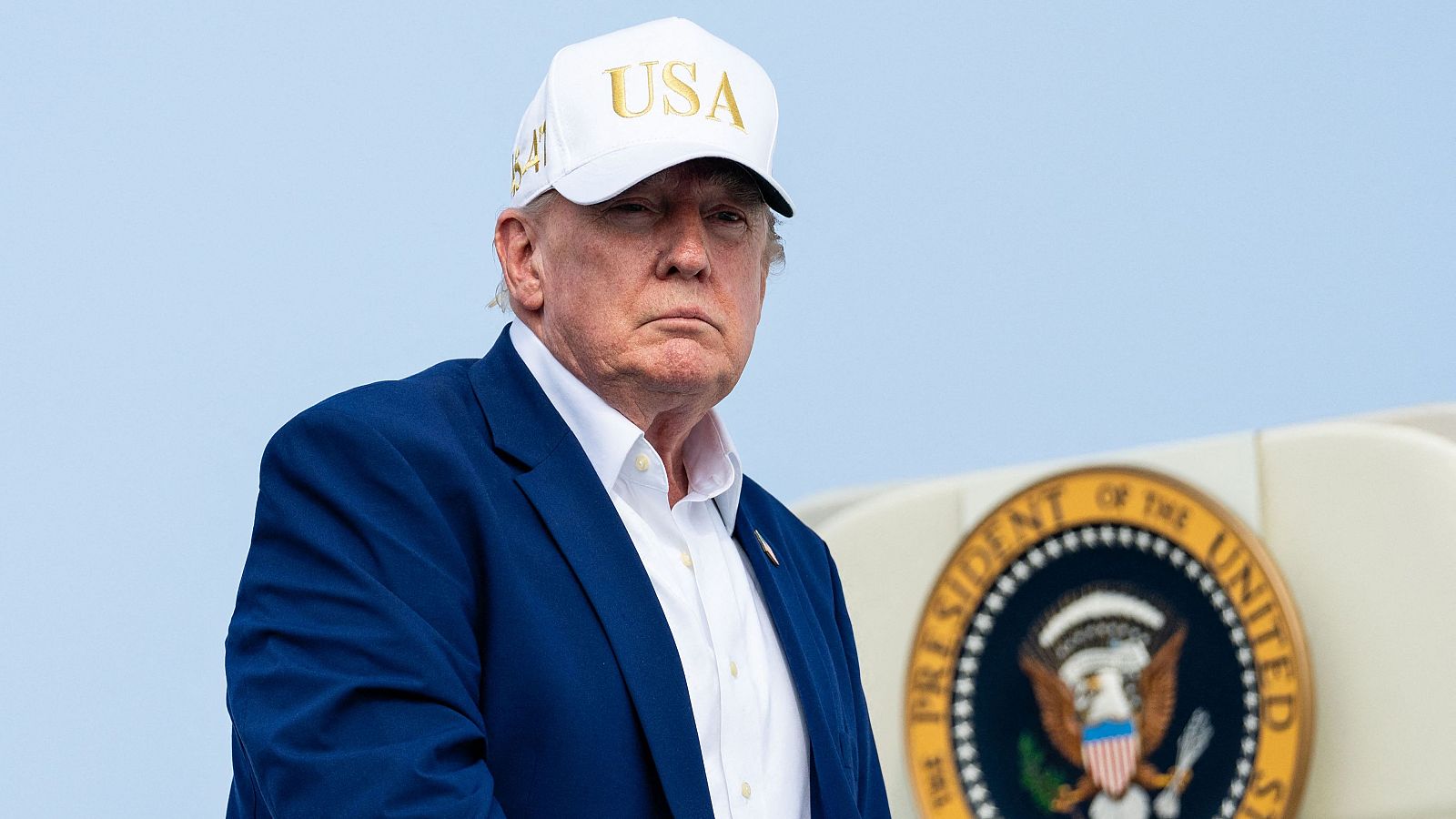 Donald Trump, con gorra "USA 45" y traje azul, posa con expresión seria frente al Sello Presidencial de EE. UU. en un día soleado.