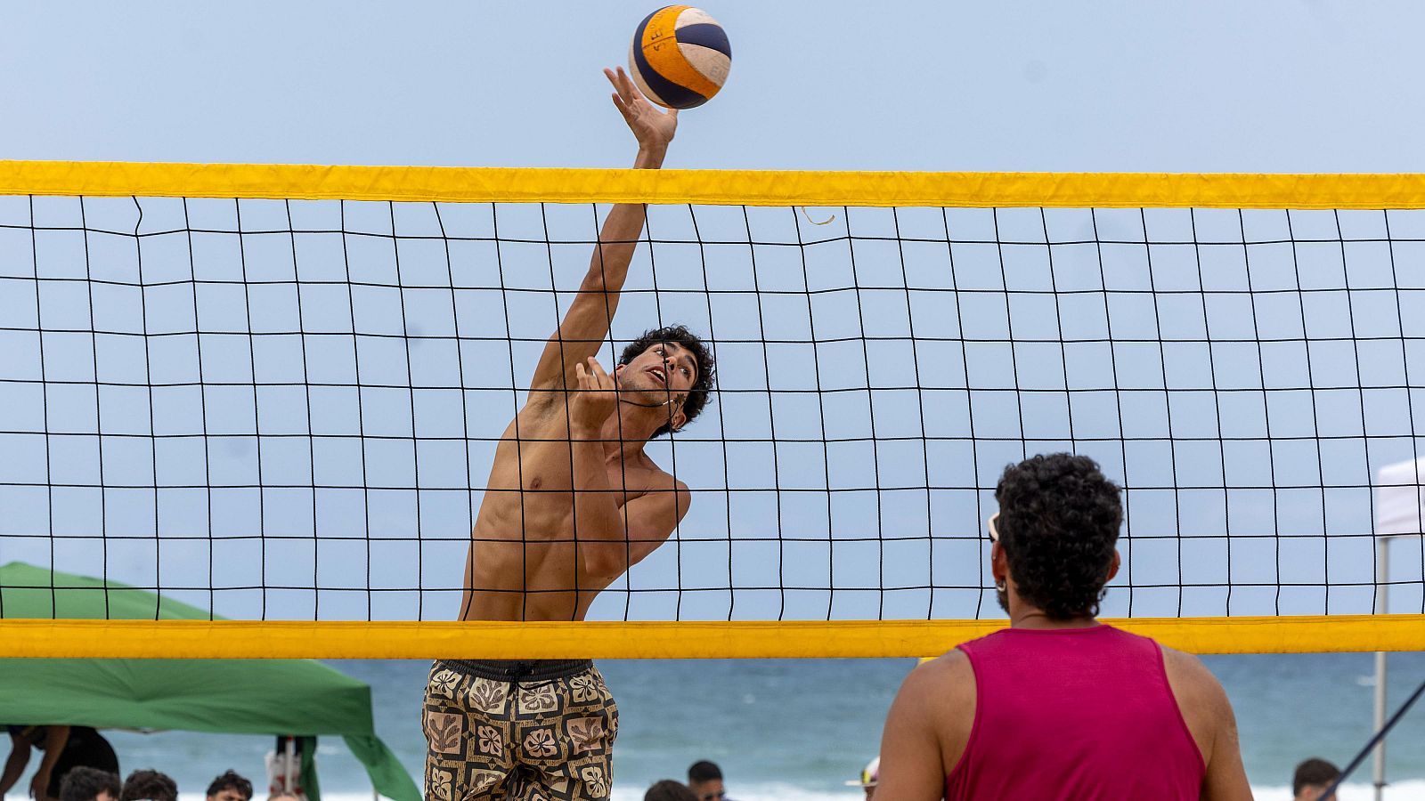 Joven jugador de voleibol de playa realiza un potente remate. Balón en el aire, red amarilla, arena y mar de fondo. Atmósfera de deporte y ocio.