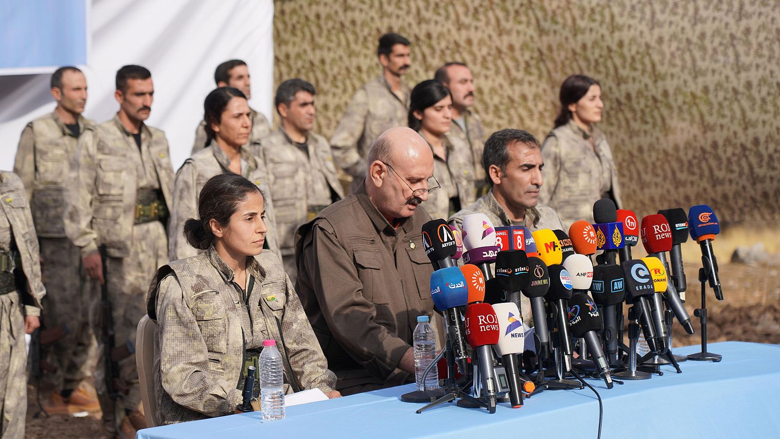 Conferencia de prensa del PKK al aire libre con combatientes uniformados. Un hombre con gafas y bigote habla rodeado de micrófonos de diversos medios como ROJ news, ANF y Rudaw.