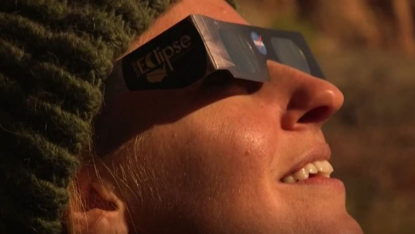 Mujer con gafas de eclipse de la NASA, perfil derecho, boca abierta, gorro verde, fondo cálido desenfocado.