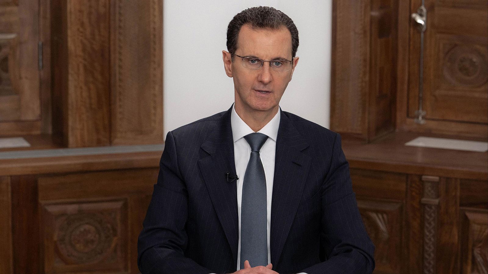 Bachar al Asad, expresidente sirio, en un discurso televisado el 16 de febrero de 2023. Viste traje oscuro, camisa blanca y corbata grisácea, con gafas y micrófono de solapa.