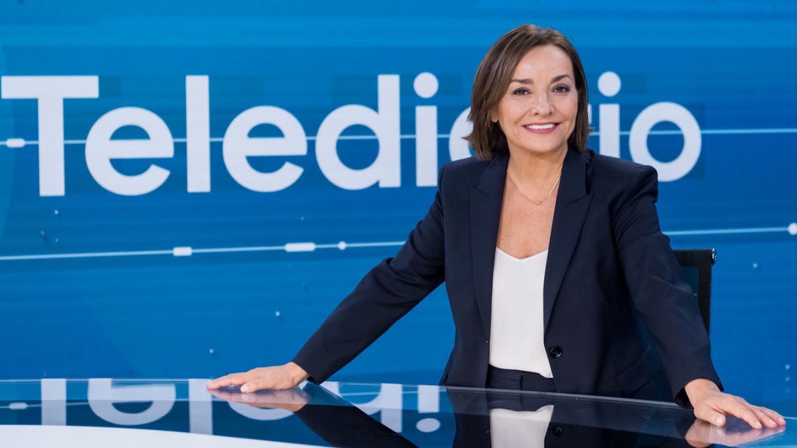 Pepa Bueno, periodista, en un plató de televisión con fondo azul y el texto "Telediario". Viste traje azul marino y blusa blanca.