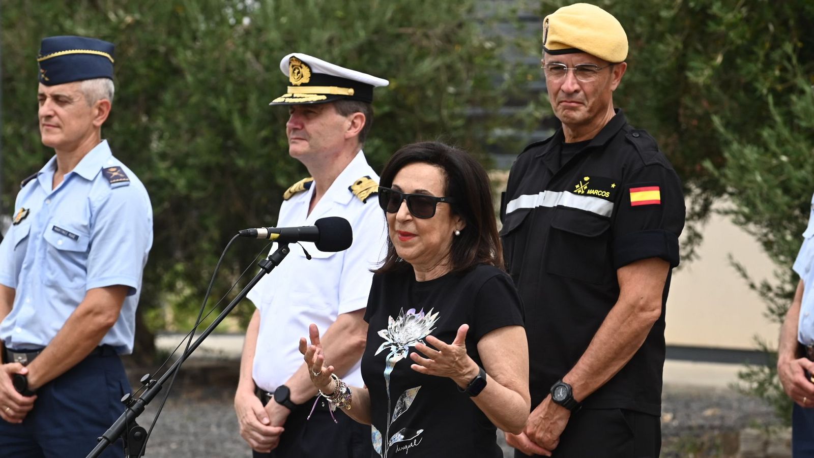 Margarita Robles, ministra de Defensa, con gafas de sol y camiseta floral, habla en un evento exterior rodeada de oficiales militares.