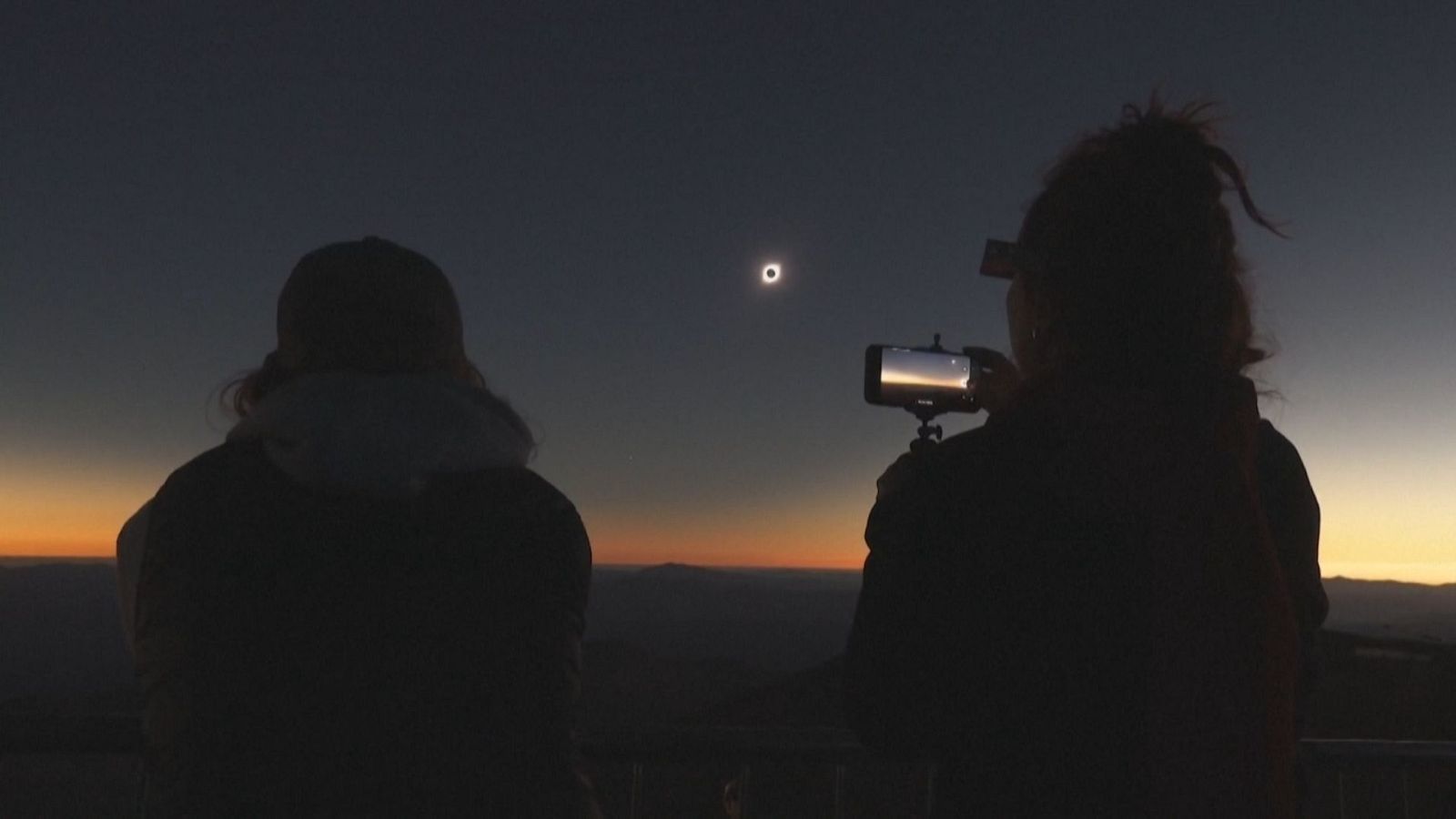 Eclipse total de sol observado por dos personas en silueta desde un mirador, con la corona solar destacando en un cielo crepuscular.