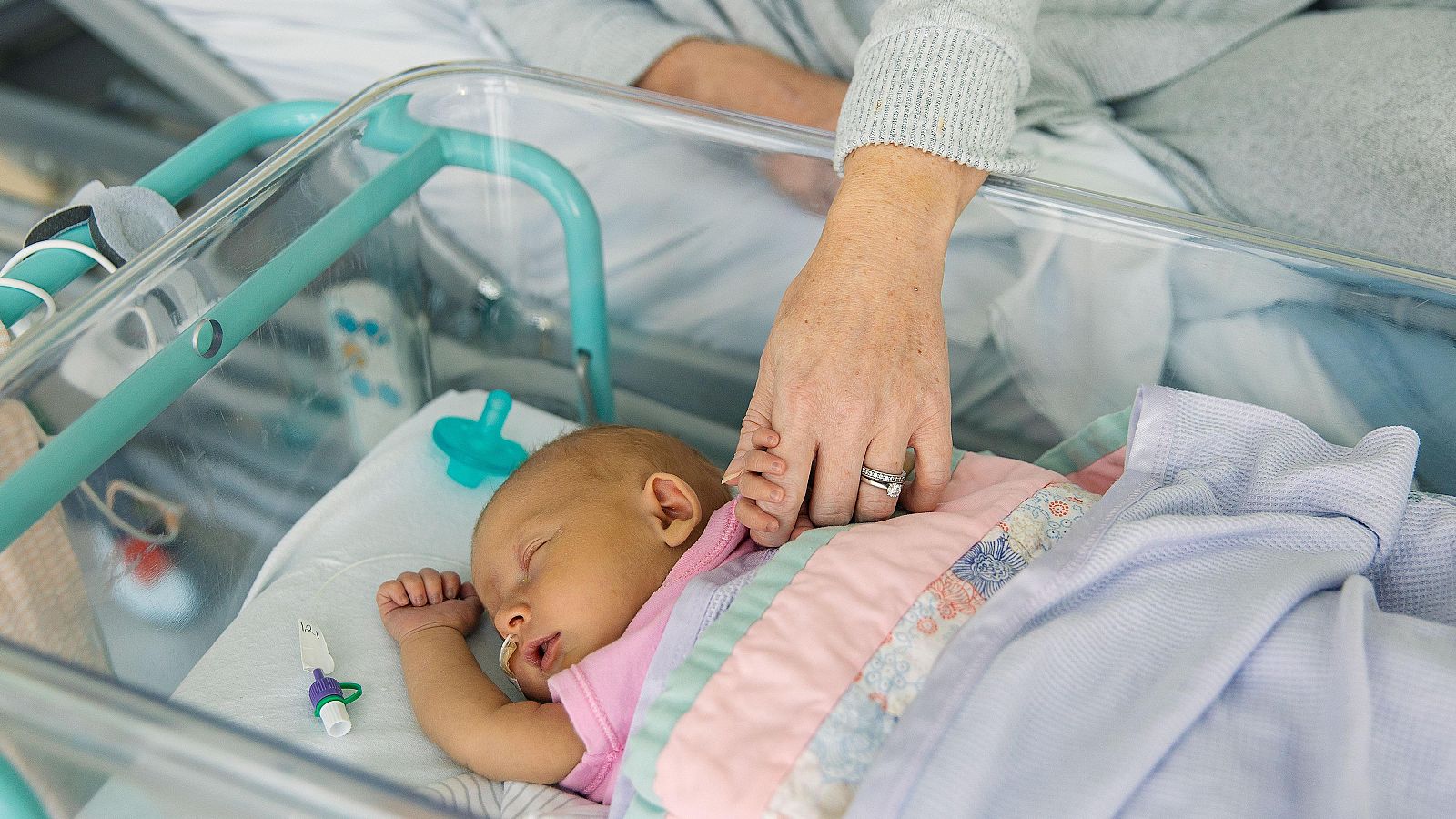Un bebé recién nacido agarra desde la cuna con sus deditos la mano de su madre tumbada en la cama.
