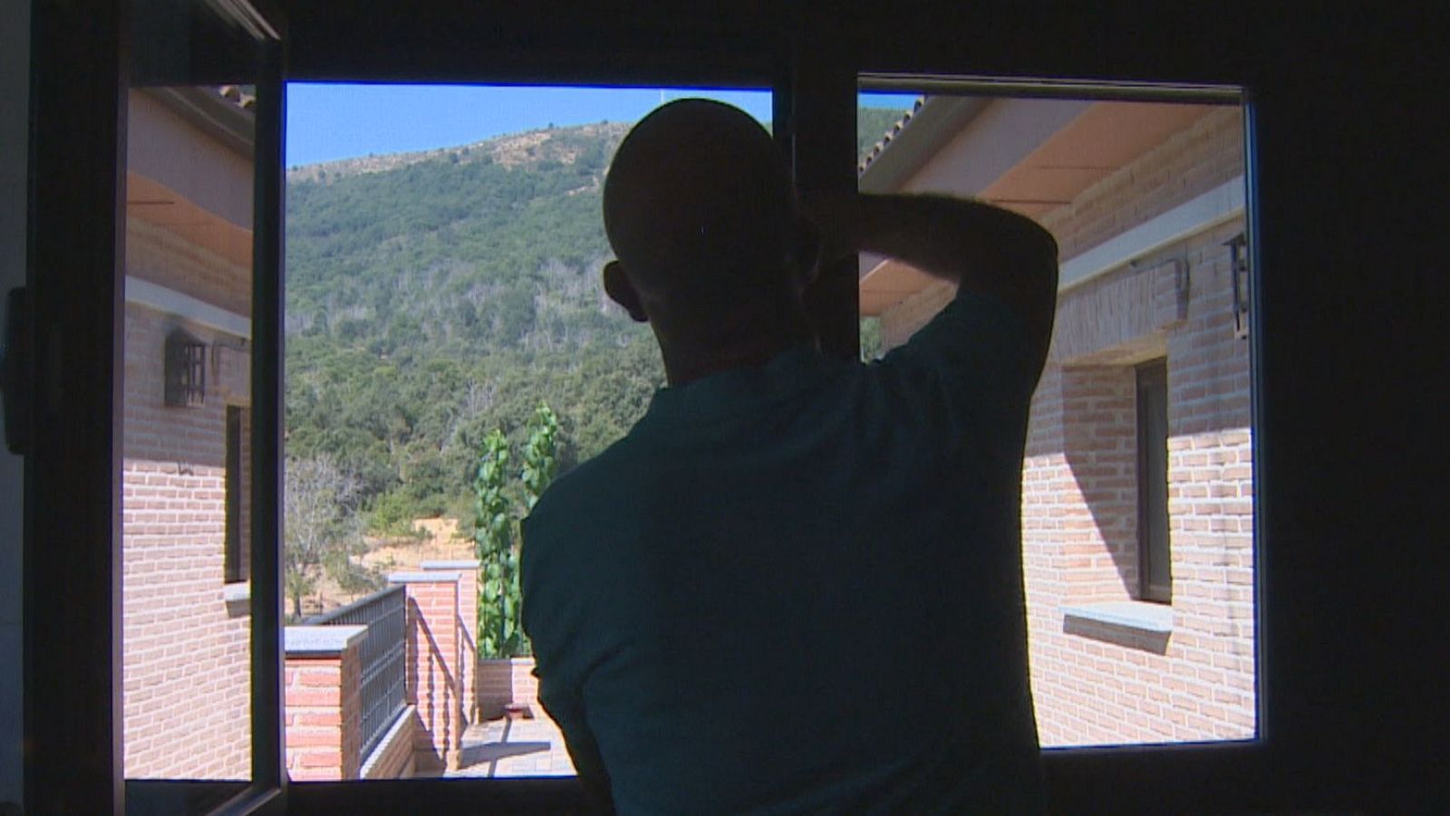 Hombre calvo en silueta observa un paisaje soleado a través de una ventana, con edificaciones y una sierra verde al fondo.