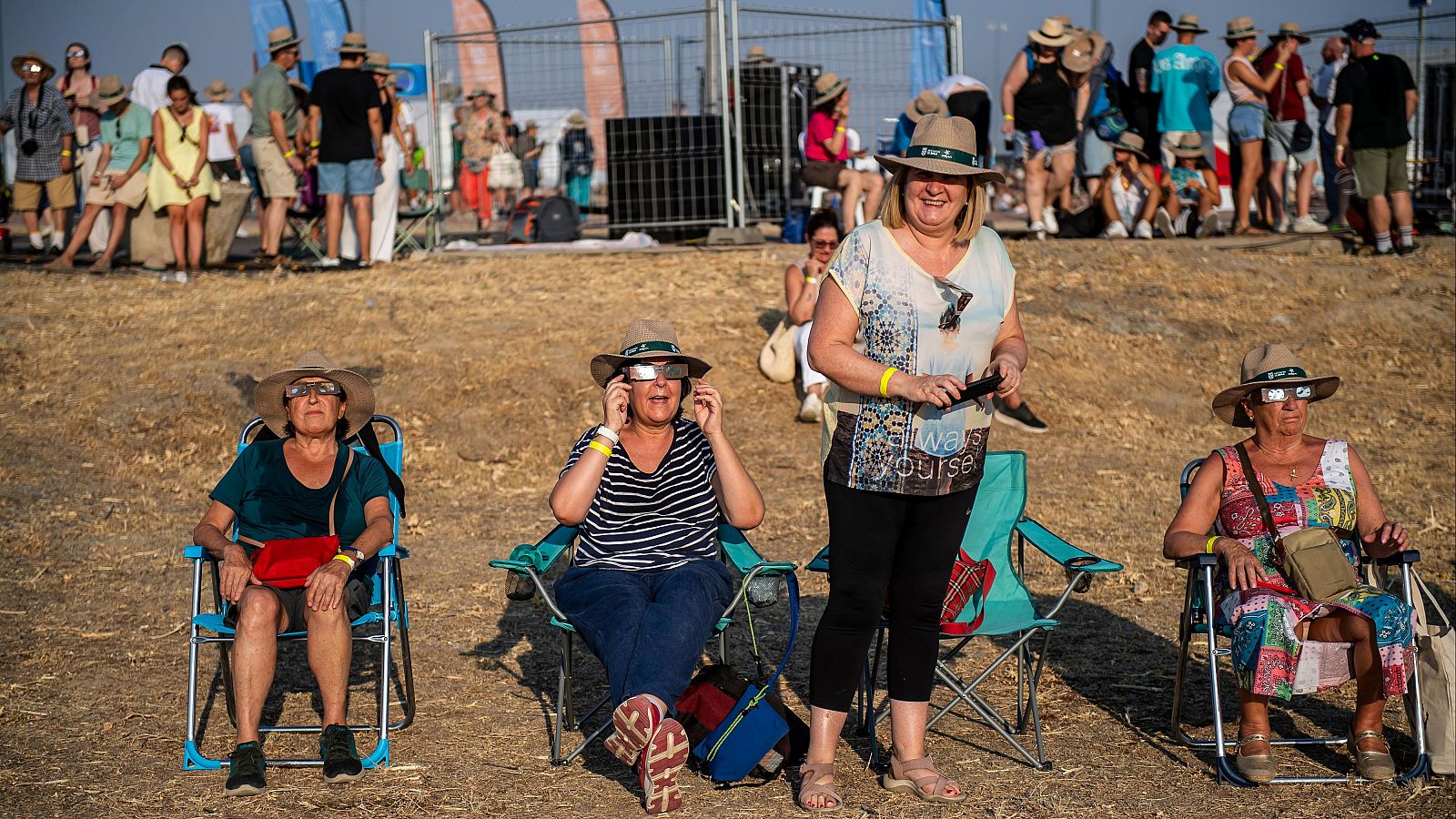 Mujeres con sombreros y gafas de eclipse observan un evento solar en un día soleado, sentadas y de pie en un terreno abierto.