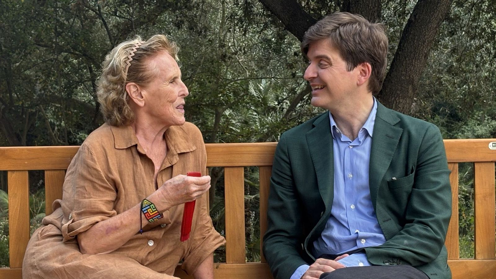 Mercedes Milá y Marc Giró conversan al aire libre. Ella, con diadema y camisa marrón, sostiene un objeto rojo. Él, con camisa azul y americana verde, sonríe.
