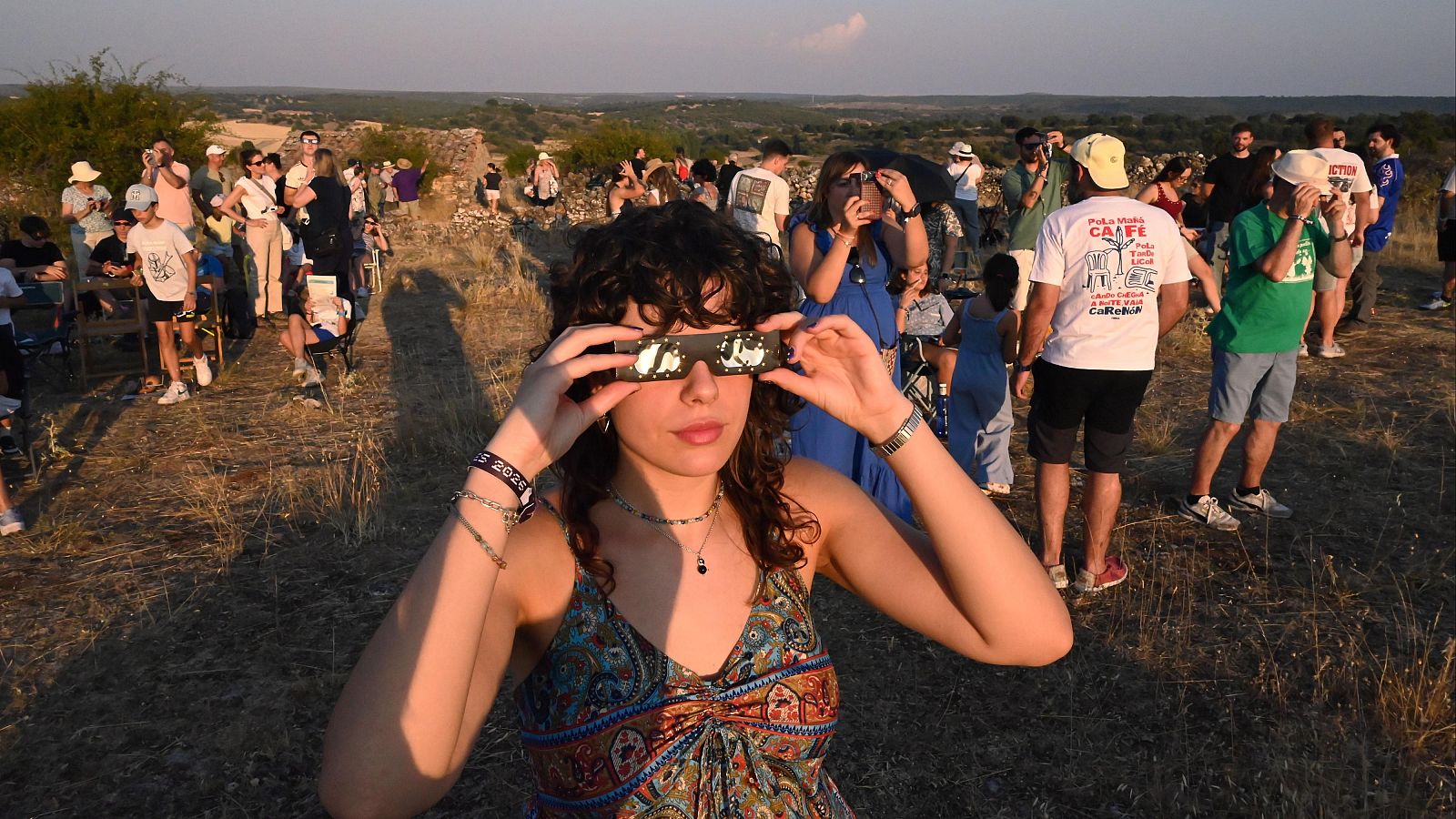 Mujer con gafas de eclipse y pulsera '2026' observa evento astronómico junto a multitud en ladera.