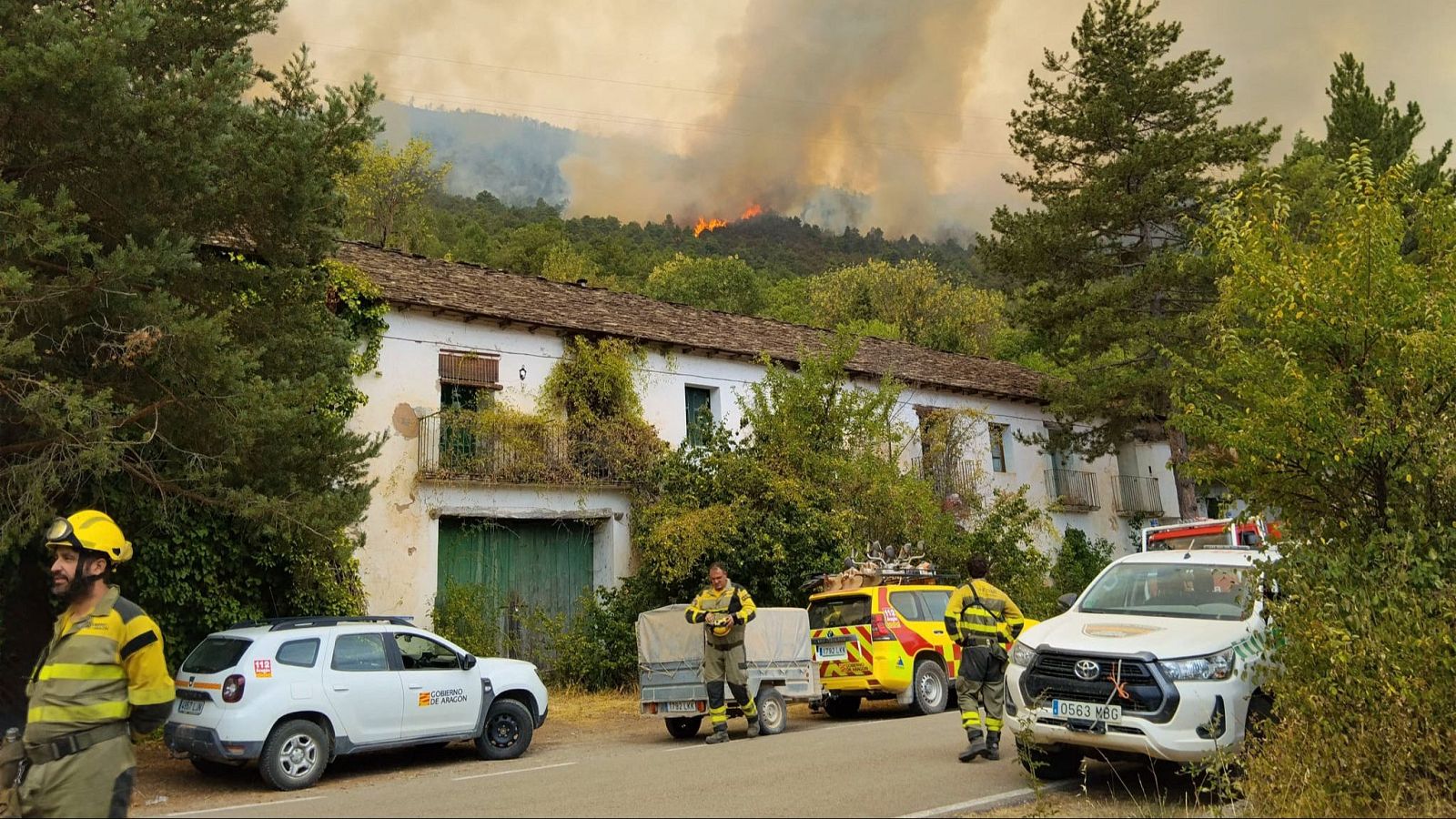 Bomberos junto a una casa con un incendio forestal activo de fondo y columnas de humo.