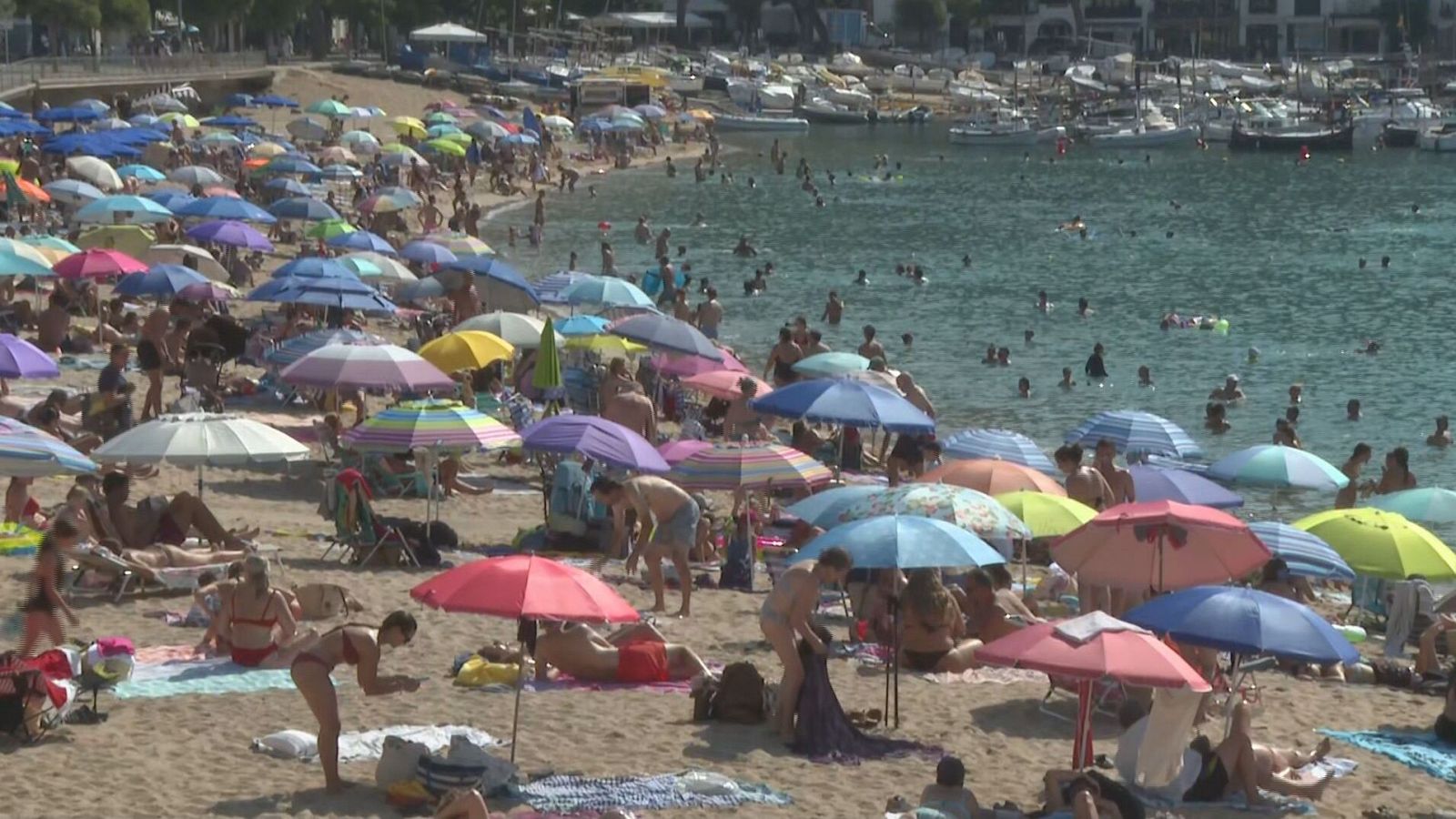 Playa abarrotada con sombrillas de colores, gente en la arena y en el mar azul verdoso. Embarcaciones de recreo y edificaciones costeras al fondo.