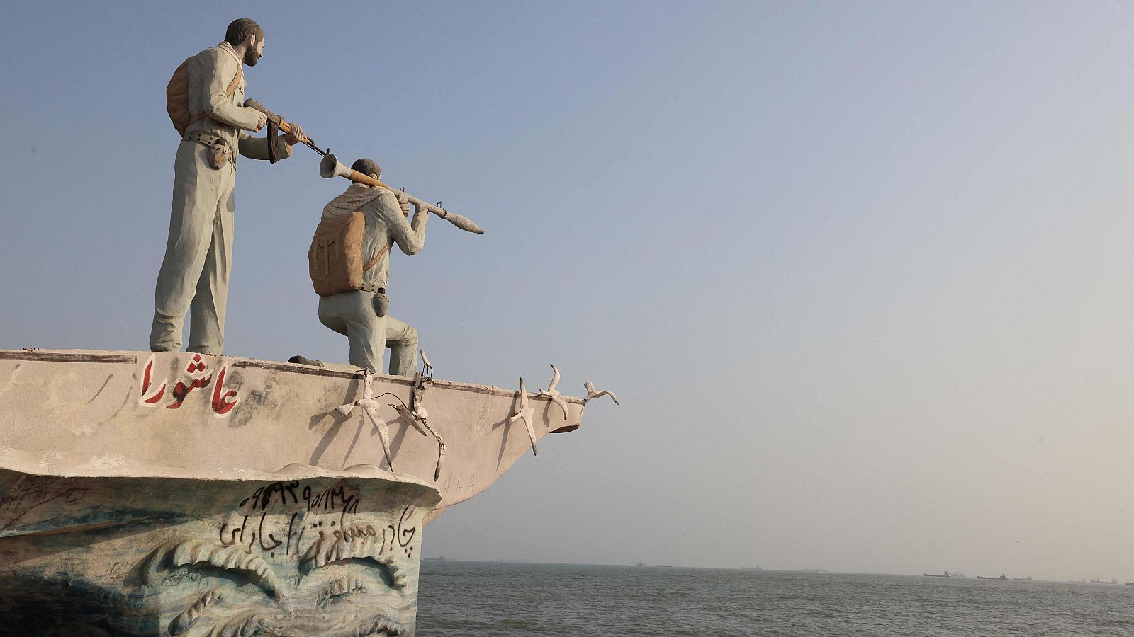 Escultura de dos combatientes con armamento, erigida sobre una proa, con inscripciones en persa/árabe "Ashura" en rojo. Vista al mar y cielo azul. Bandar Abás, Irán, 10 de agosto de 2026.