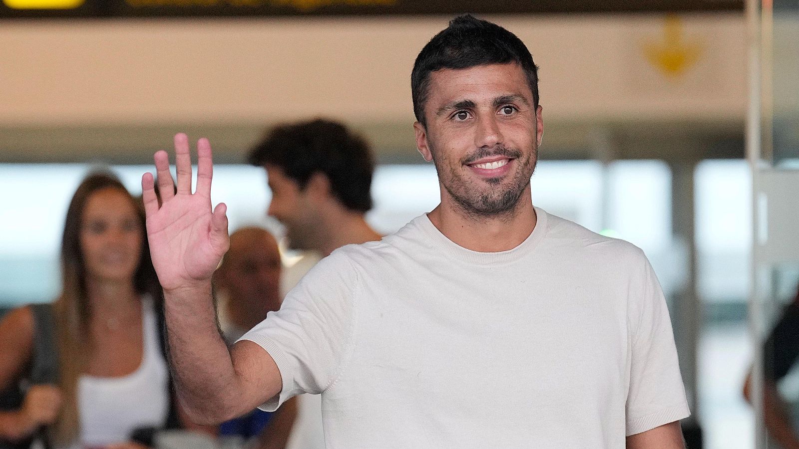 Rodri saluda a su llegada a Barcelona, sonriendo y con el brazo levantado en un gesto amistoso. Se le ve en un aeropuerto, vestido con una camiseta beige.