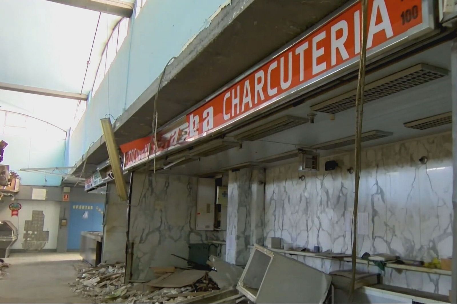 Interior de un mercado municipal abandonado, con escombros, restos de puestos y un cartel de "CHARCUTERIA 100".