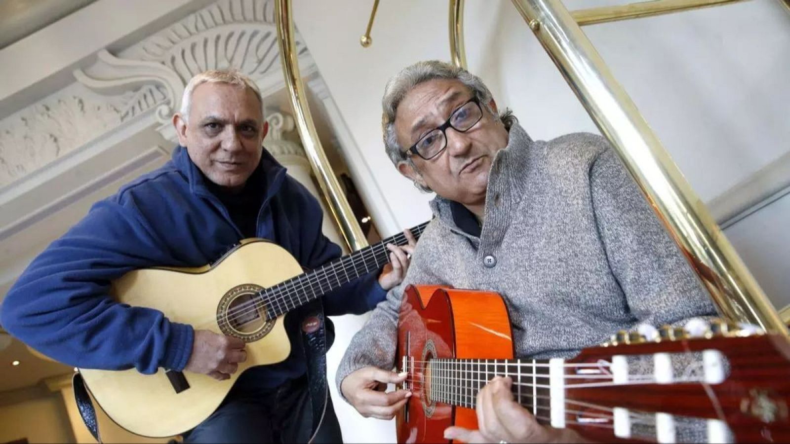 José y Delfín Amaya, del dúo "Los Amaya", posan con sus guitarras acústicas en un interior. José (i) con chaqueta azul y guitarra clara; Delfín (d) con gafas, cárdigan gris y guitarra rojiza.