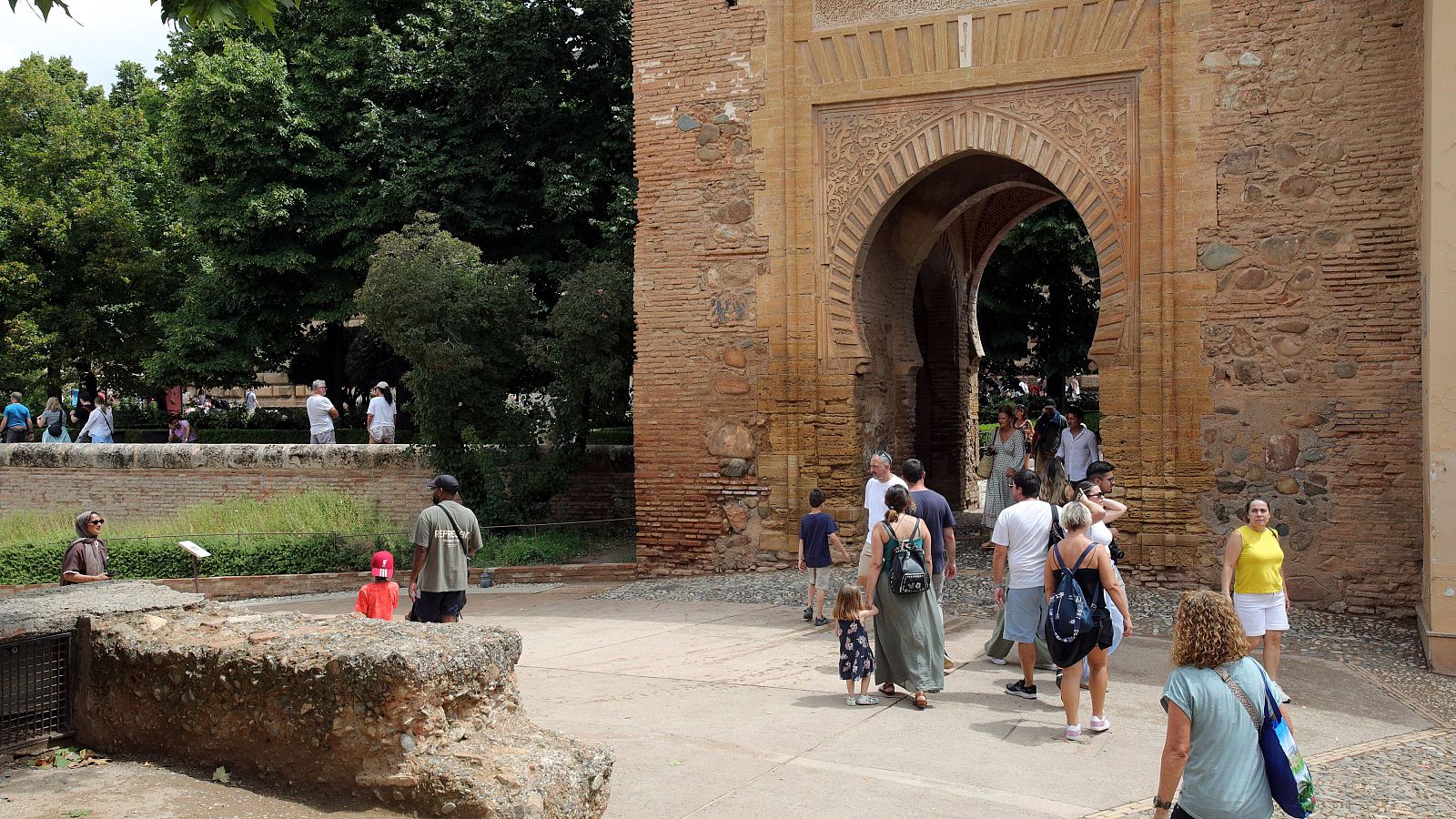 Arco nazarí en la Alhambra con turistas y vegetación exuberante. Se observan restos de estructuras antiguas en primer plano.