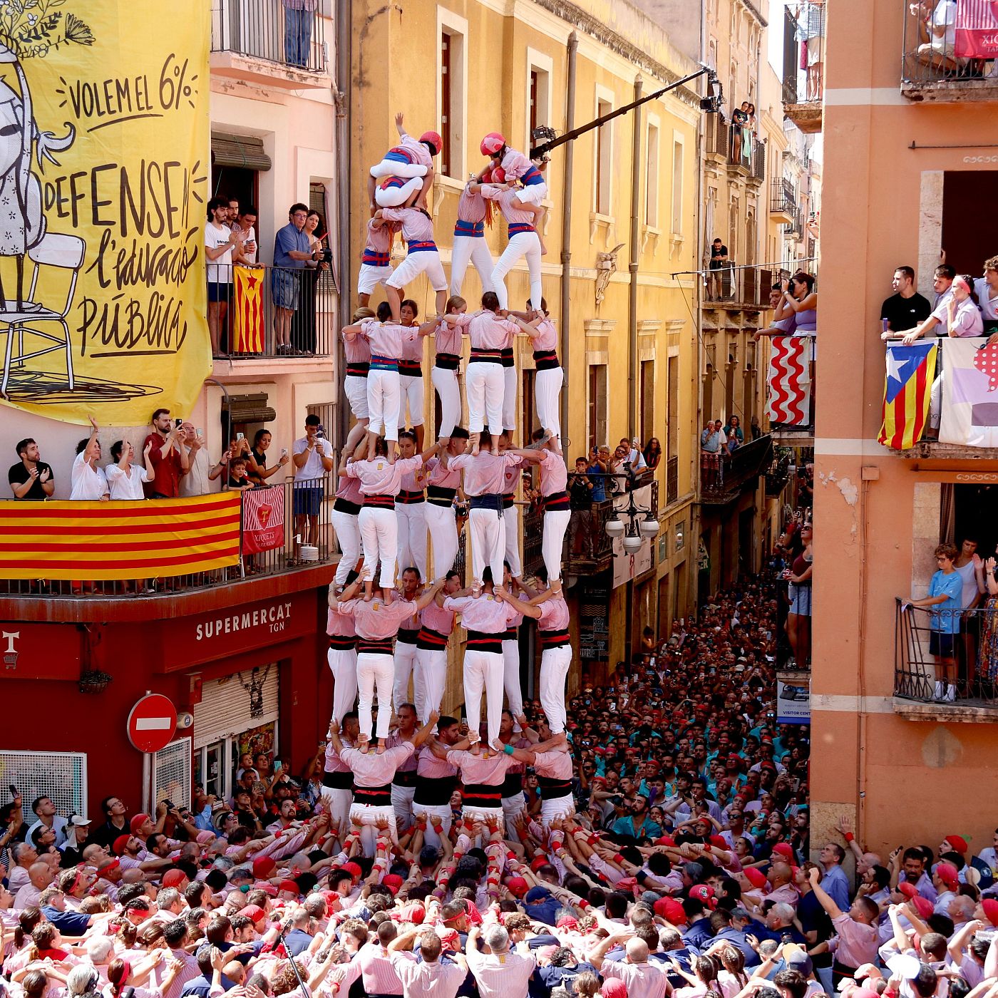 Torre humana "3 de 9 amb folre" con "castellers" de rosa y blanco, culminada por un "enxaneta". Pancarta "VOLEM EL 6% DEFENSA L'EDUCACIÓ PÚBLICA" y banderas catalanas visibles. Multitud en la "pinya".