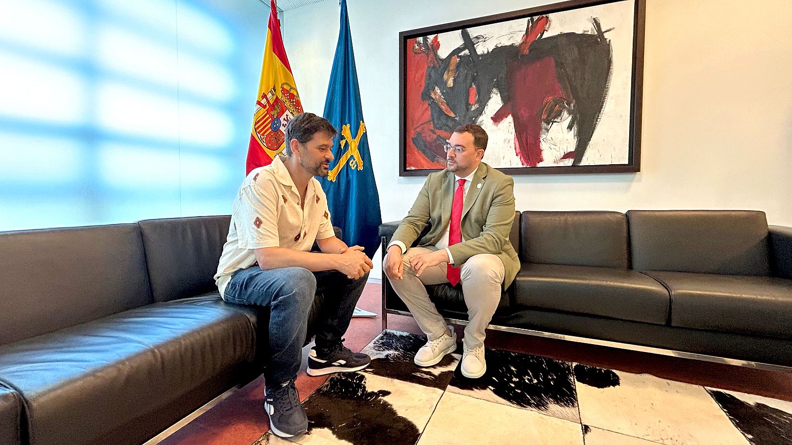 Adrían Barbón y Pablo Martínez Corral conversan sentados en sofás en una sala institucional, con las banderas de España y Asturias al fondo.