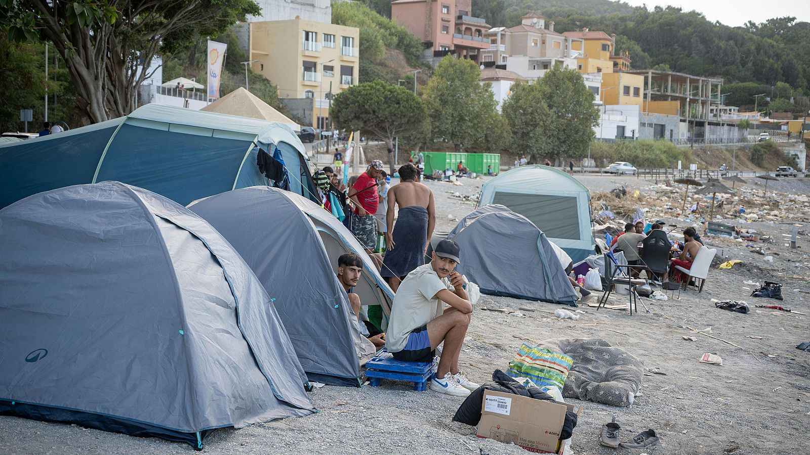 Tiendas de campaña improvisadas en la playa del Trampolín de Ceuta, con grupos de jóvenes migrantes sentados.