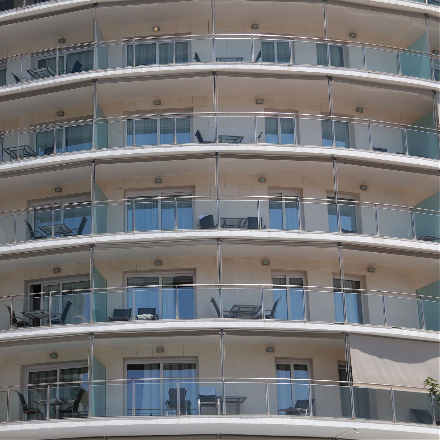Edifici modern amb façana corba, balcons amb baranes de vidre i mobiliari d'exterior. Es distingeix vegetació a la cantonada inferior dreta.