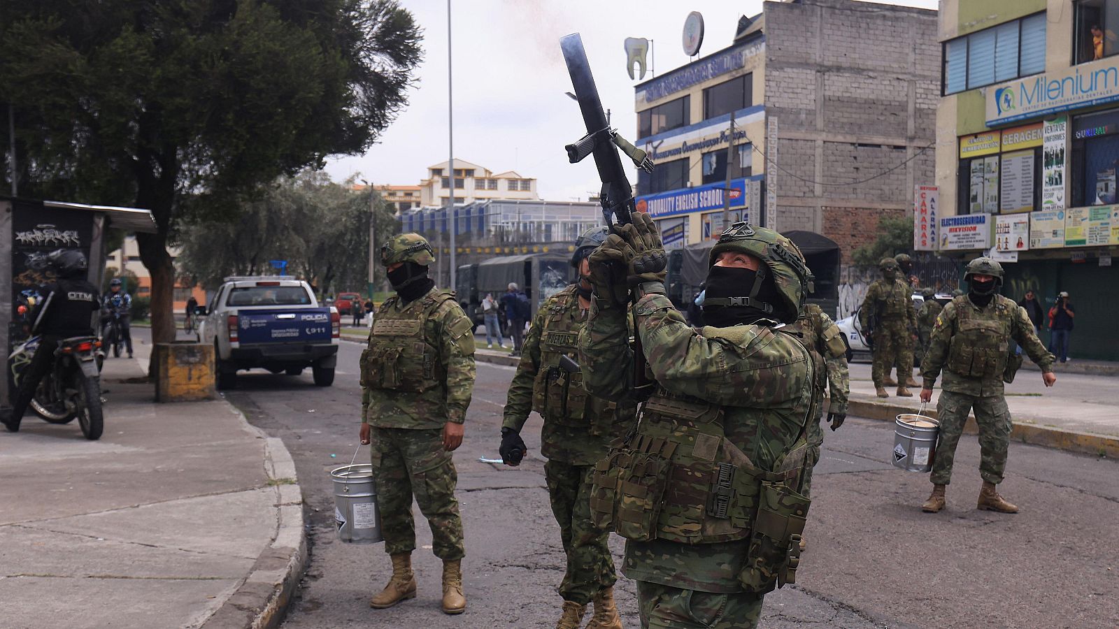 Militares ecuatorianos en acción urbana, uno dispara un lanzador con humo rojizo. Se ven vehículos policiales y edificios con letreros como 'QUALITY ENGLISH SCHOOL' y 'Milenium CLINICAS'.