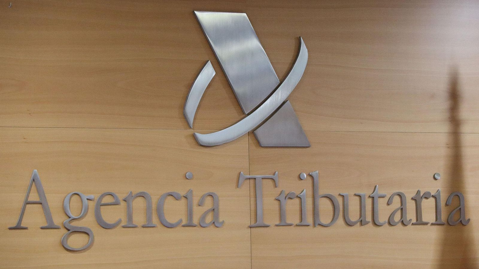 Logotipo y nombre "Agencia Tributaria" en metal plateado con relieve sobre madera clara. La "A" y la "T" están en mayúscula.