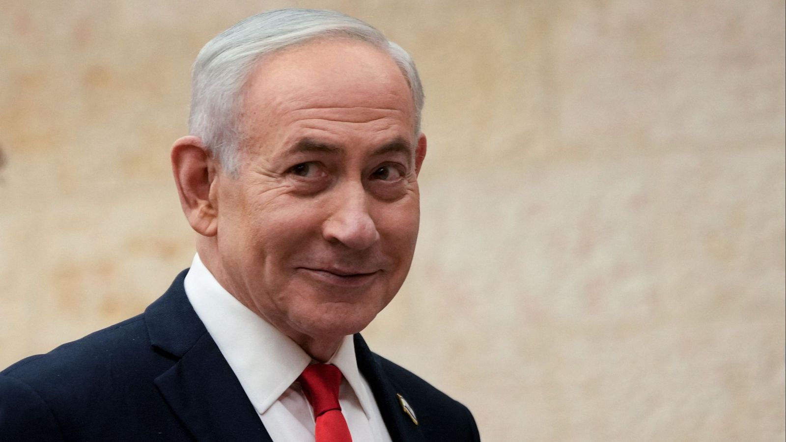 Benjamin Netanyahu, político israelí, en un primer plano con traje oscuro, camisa blanca y corbata roja. Cabello canoso, ligera sonrisa, fondo liso y desenfocado.