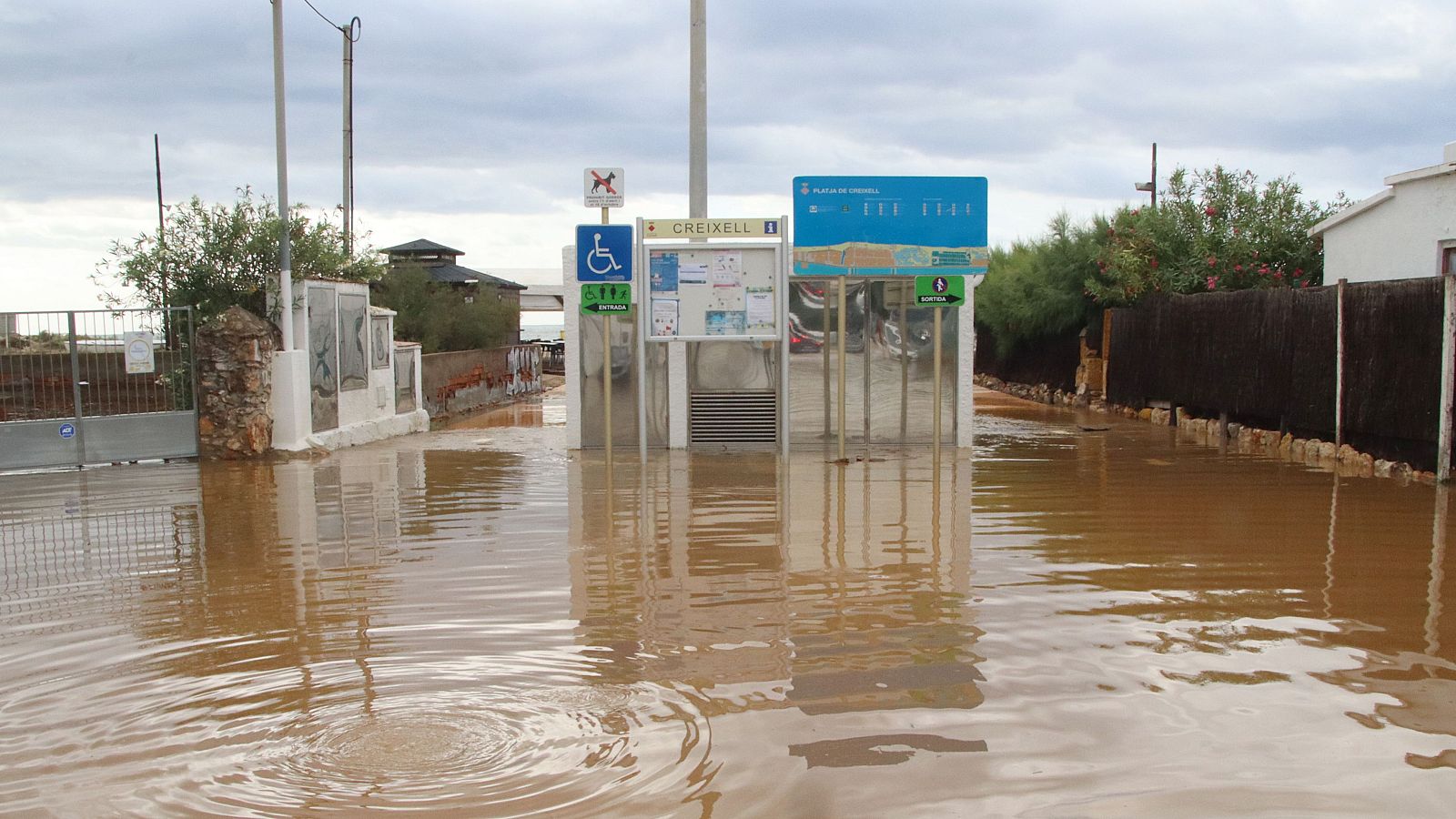 Inundación en los accesos a la playa de Creixell, Baix Penedès. Agua turbia cubre la zona, reflejando estructuras. Señales de accesibilidad, aseos y nombre de la playa visibles.