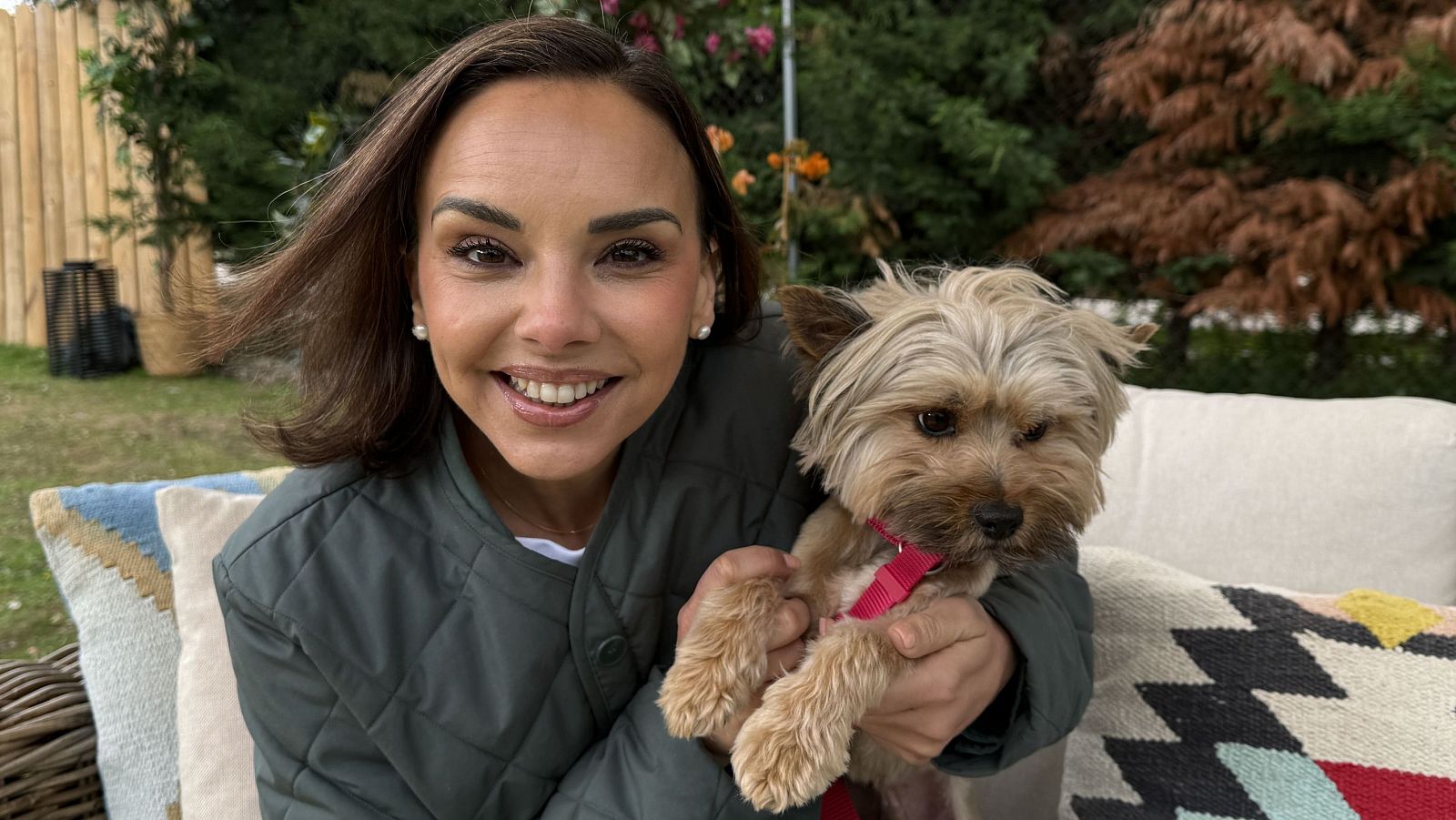 Raquel Sánchez Silva sonríe a la cámara mientras abraza a un Yorkshire Terrier con arnés rosa, sentados en un sofá de exterior con cojines.