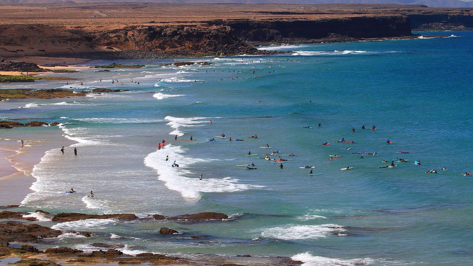 Vista aérea de una costa con playa, mar turquesa y decenas de personas practicando surf entre olas y formaciones rocosas, con acantilados áridos al fondo.